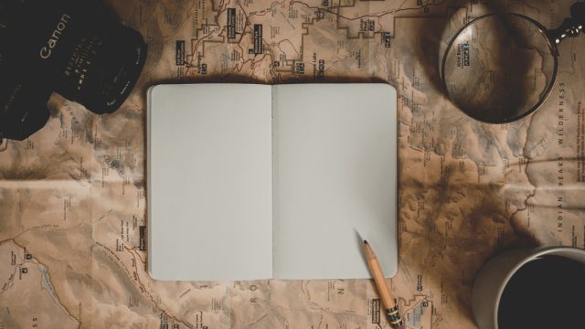 open journal with blank pages centered on brown and white map in between a canon camera and a magnifying glass